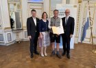 Gruppenfoto im Spiegelsaal der Regierung der Oberpfalz: Eine Frau und ein Mann in Tracht halten die Urkunde des Integrationspreises 2025. Neben ihnen stehen Staatssekretär Sandro Kirchner und Regierungspräsident Walter Jonas. Im Hintergrund ist ein Banner mit der Aufschrift „Integration leben – In Bayern daheim“ sowie eine bayerische Flagge zu sehen.